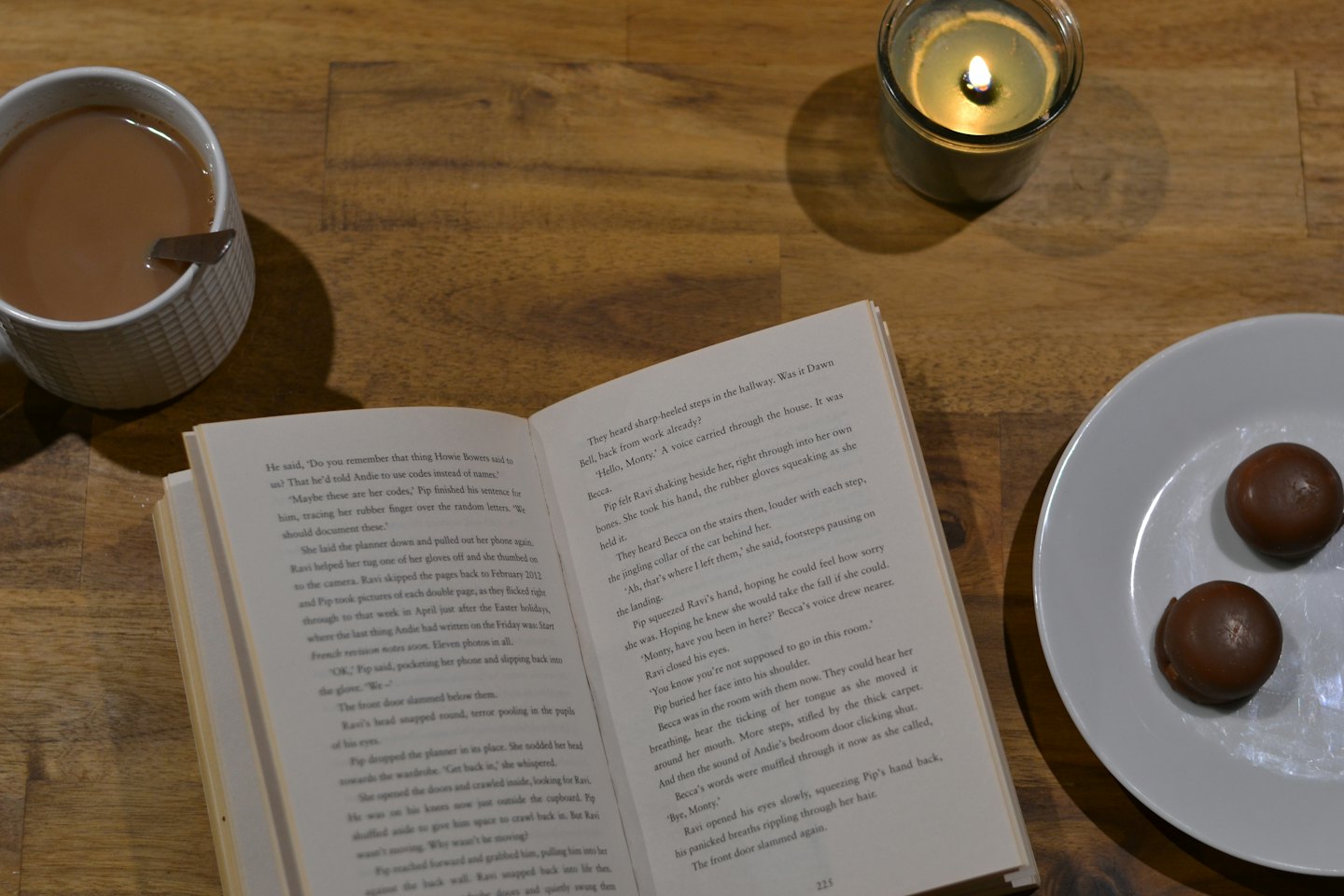 An open book, tea, candle, and chocolate on a table.