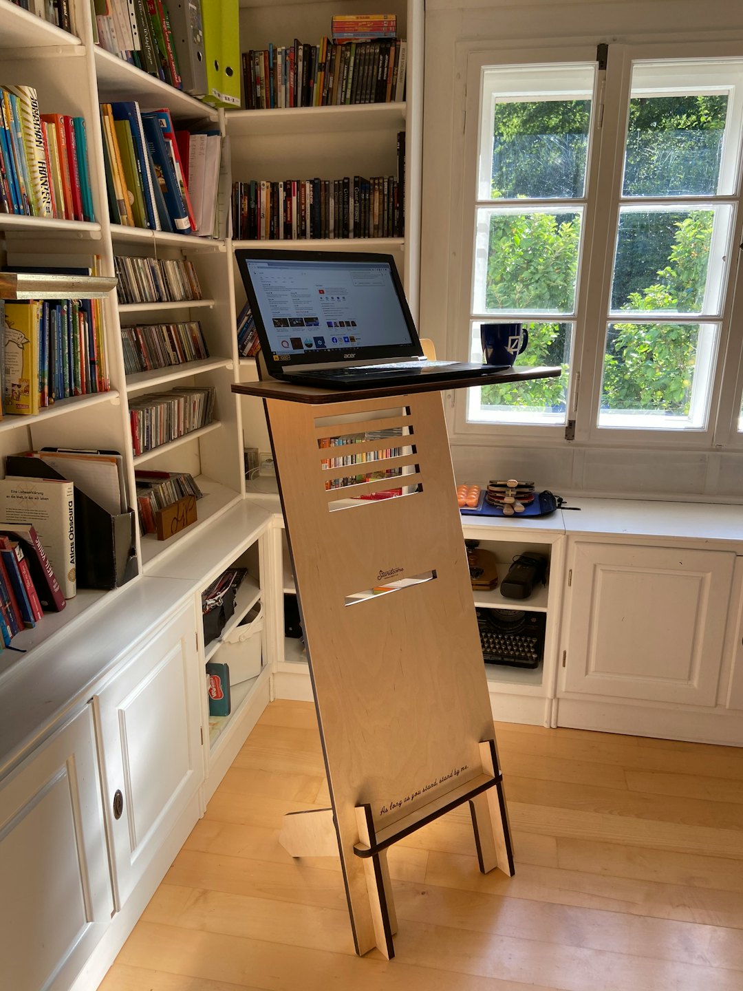 Standing desk with laptop and blue mug by sunlit window.